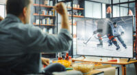 At Home Ice Hockey Fans Sitting on a Couch Watch Game on TV, Cheer when Favourtite Sports Team to Win the Championship. Screen Shows Professional Players During World Cup. Over the Shoulder