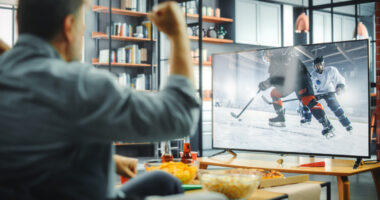 At Home Ice Hockey Fans Sitting on a Couch Watch Game on TV, Cheer when Favourtite Sports Team to Win the Championship. Screen Shows Professional Players During World Cup. Over the Shoulder