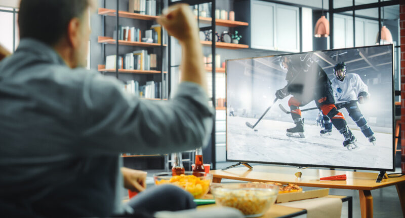 At Home Ice Hockey Fans Sitting on a Couch Watch Game on TV, Cheer when Favourtite Sports Team to Win the Championship. Screen Shows Professional Players During World Cup. Over the Shoulder