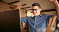 Handsome businessman with laptop having his arms with fists raised, celebrating success. Happy freelancer hipster in glasses finished work on project. Man won a lot of money in lottery prize